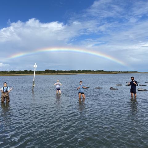 Students stand in the ocean with a rainbow in the background