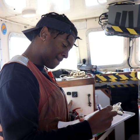 A student researcher on a ship takes notes on a clipboard 