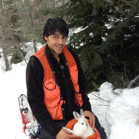 Alex Kumar kneeling in a snowy forest with a white snowshoe hare in his lap.