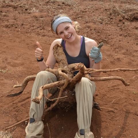 A young woman holds a large root in her lap and gives thumbs up