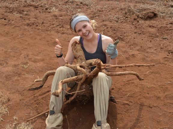 A young woman holds a large root in her lap and gives thumbs up